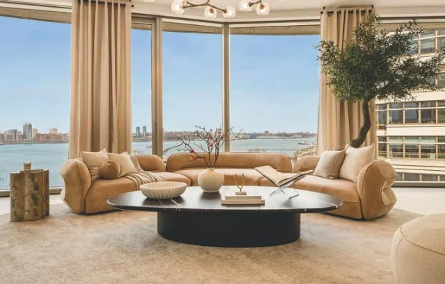 A warm living room with curved sofas, wide windows, a Hudson River view and Manhattan buildings beyond.