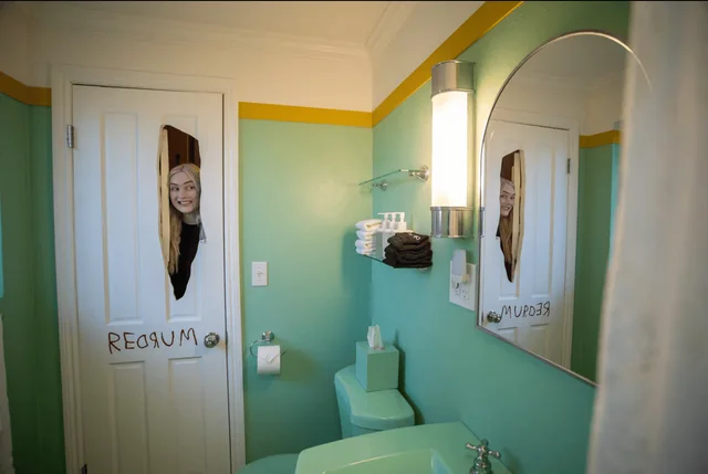 A bathroom with mint-green walls and yellow trim, a white door with an axe-shaped opening through which a woman's face appears, and the word REDRUM written across the lower panels