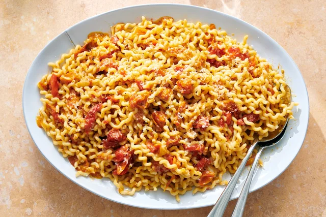 An overhead photograph of a wide white bowl of long fusilli tossed with a sauce of torn roasted tomatoes, a scatter of grated Parmesan over the top, and two serving spoons resting at the right edge