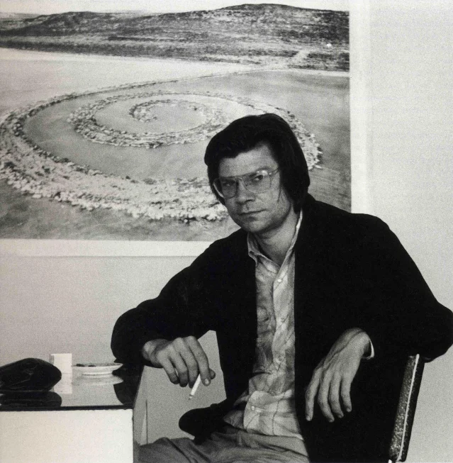 Robert Smithson 坐在桌旁抽烟，身后的墙面悬挂着《Spiral Jetty》的图像。