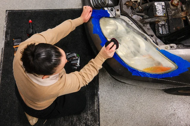 Seen from above, a person kneeling at the front corner of a car with the hood open; blue painter's tape outlines the hazy plastic headlight lens, and one hand presses a small black polishing pad against the lens, with the battery and engine bay visible at the top of the frame