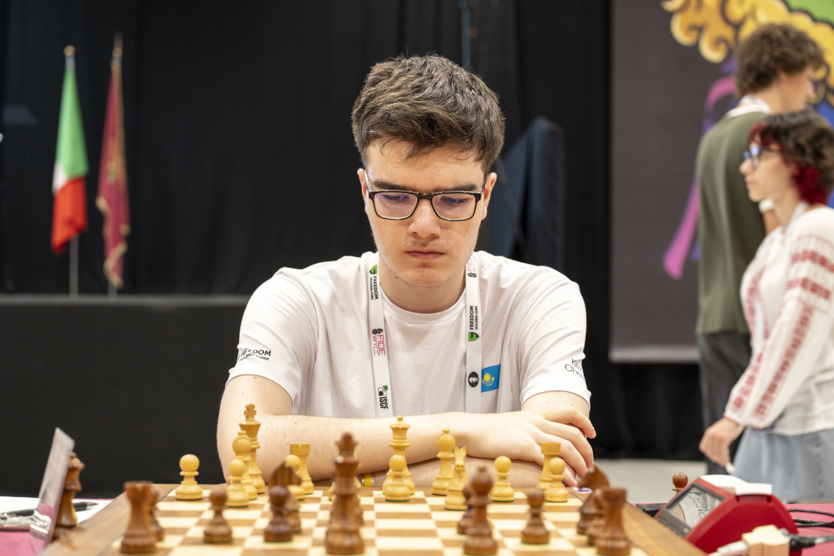 A young player in glasses and a white shirt at a chessboard, flags behind him