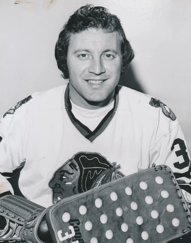 A black-and-white portrait of a smiling man in a light hockey jersey with a dark shoulder yoke and a large circular crest, holding a goalie stick with a perforated blade across his chest