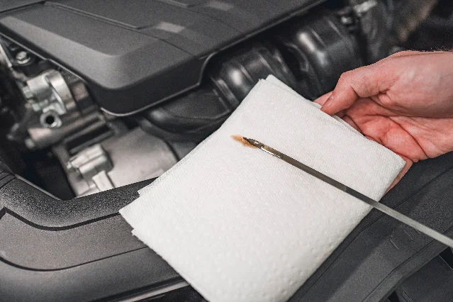 A metal oil dipstick lying on a white paper towel held in front of an open engine bay