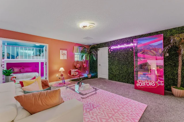 A pink Barbie-themed lounge with a transparent Barbie Box photo booth, flamingo chairs, palms, and a neon room sign
