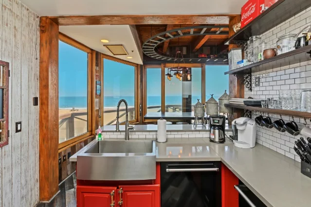 A galley kitchen with a farmhouse sink, red cabinets and tile, windows onto the ocean, and a model train track curving across the ceiling above