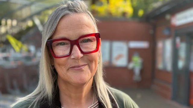a woman with grey-blonde hair and red-framed glasses standing outside a red-brick railway station