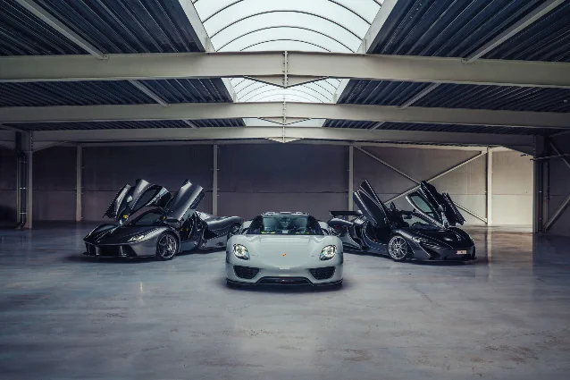 Three hypercars lined up in an industrial hall with a glass skylight ceiling: a black Ferrari LaFerrari with its doors open, a grey Porsche 918 Spyder, and a black McLaren P1 with its doors open