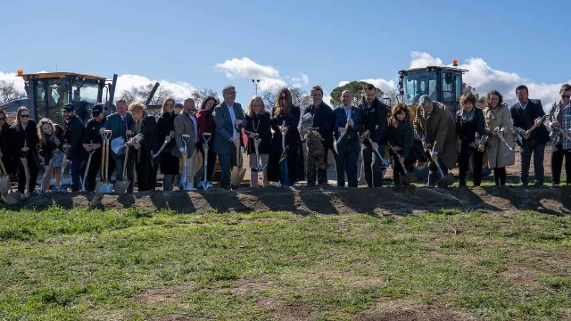 A line of people holding shovels on a bare dirt site, with two pieces of earth-moving equipment behind them
