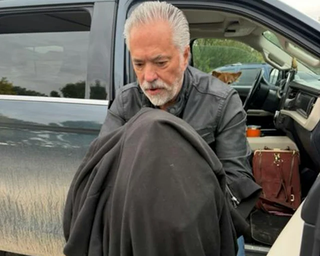 A man in a grey jacket bending over a dark bundle held against his chest beside an open car door