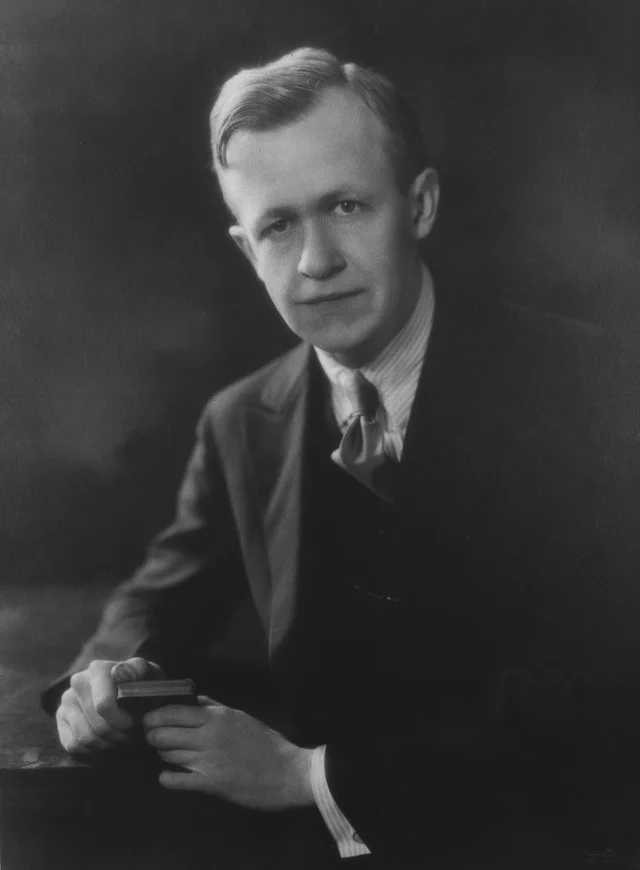 A black-and-white studio portrait of a young man in a suit and tie, one hand resting on a small book