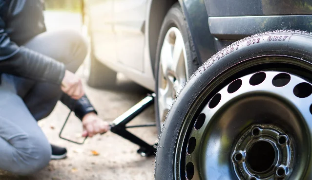 A person crouching on gravel beside a car, working the handle of a scissor jack under the sill, with a black temporary space-saver wheel standing in the foreground