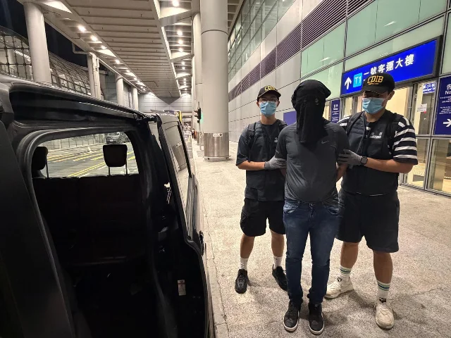 Two Hong Kong Customs officers in plain clothes escort a masked suspect outside HKIA Terminal 1 at night, with a black vehicle door open in the foreground