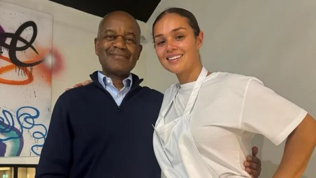 An older man in a dark blue zip-up sweater beside a smiling young woman in a white apron, with colourful artwork on the wall behind them