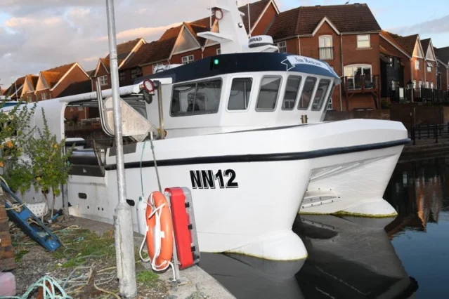 White catamaran-style fishing vessel marked NN12 moored in a marina, residential brick buildings and calm water behind it, an orange life-ring mounted on the dock post in front