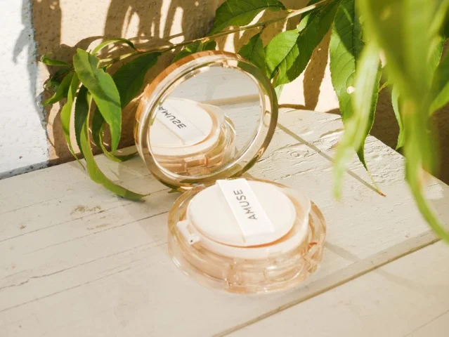 Amuse cushion compact open with puff applicator beside green leaves on a wooden surface