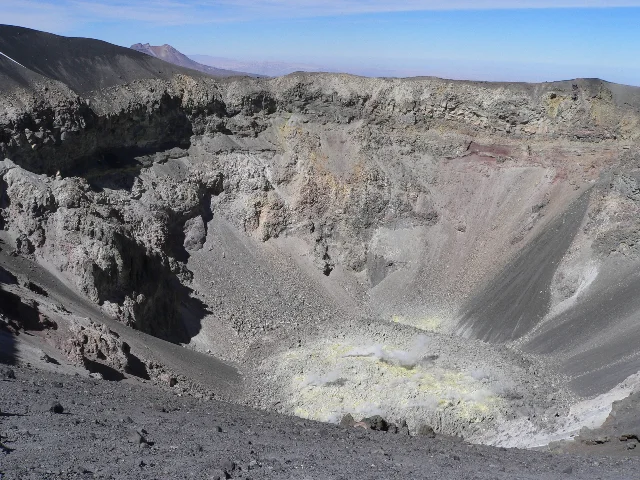 A deep grey circular crater with yellow-stained walls and a white plume of vapour rising from its floor