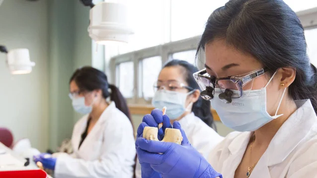 Three dental students and an instructor work with dental models and loupes in a university clinical training facility.