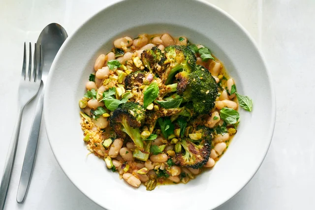 An overhead photograph of a wide pale bowl holding white beans in a golden oil, topped with charred broccoli slices, chopped pistachios, torn green herbs and crisp bread crumbs, with a silver fork and spoon at the left edge