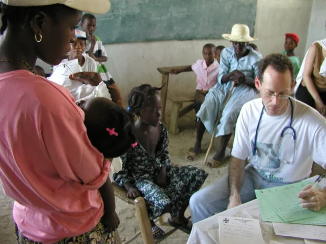 A doctor writes prescriptions at a table in a schoolroom while patients wait around him