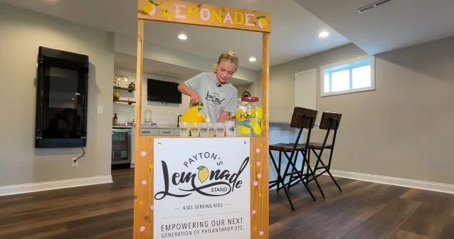 a girl at a wooden lemonade stand pouring from a yellow jug into cups, with a painted sign on the front