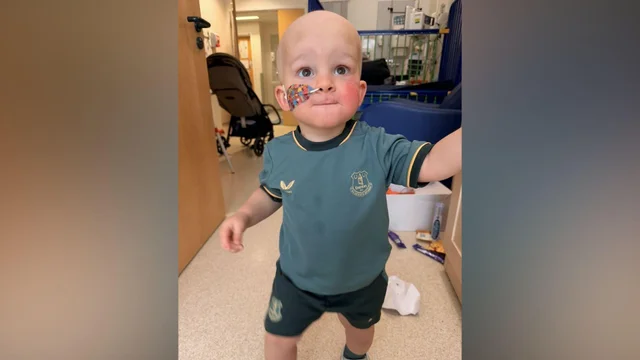 a toddler in a dark green football kit standing on a hospital corridor floor