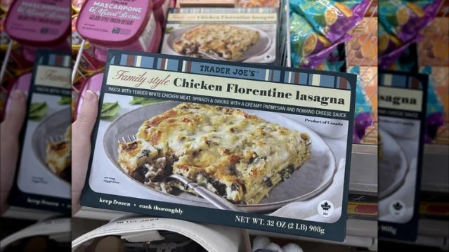 A hand holds a Trader Joe's Family Style Chicken Florentine Lasagna box in a store aisle; the front shows a cheesy lasagna portion, Family Style labeling, and net weight 32 oz