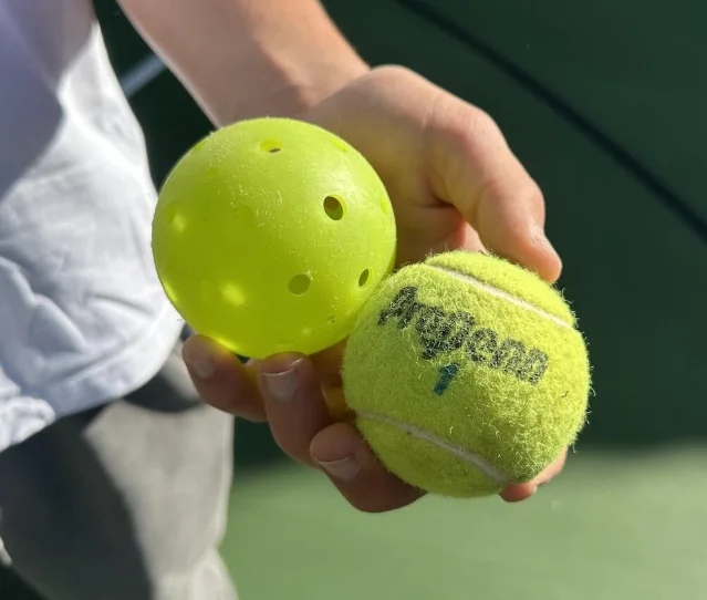 A used yellow pickleball and a worn tennis ball held side by side in a hand