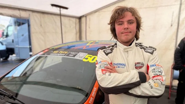 a teenager in a white racing suit with his arms folded beside an orange and blue racing car