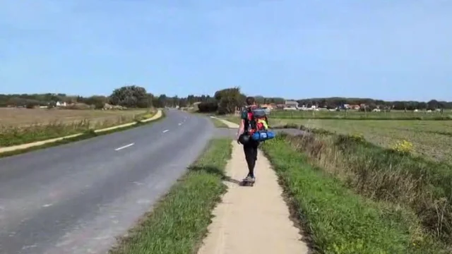 A skateboarder carrying a heavy pack pushes along a footpath beside an empty road in flat farmland under a clear sky