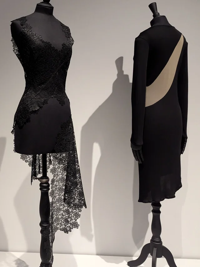 Two black outfits on mannequins, one an asymmetric lace dress, the other a long-sleeved dress with a curved opening that shows a cream lining