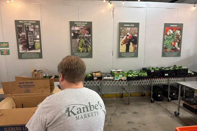 A volunteer in a Kanbe's Markets shirt in a warehouse, with four posters on the wall behind reading Healthy Corner Stores, Hunger Relief Partners, Animal Feed and Compost