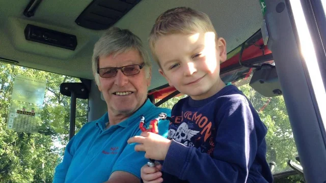 A grey-haired man in glasses and a blue shirt sitting in a tractor cab with a small blond boy on his lap