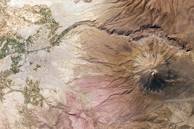 An aerial view from orbit of a grey city with a road grid on the left and a large brown volcano with dark radial ridges on the right