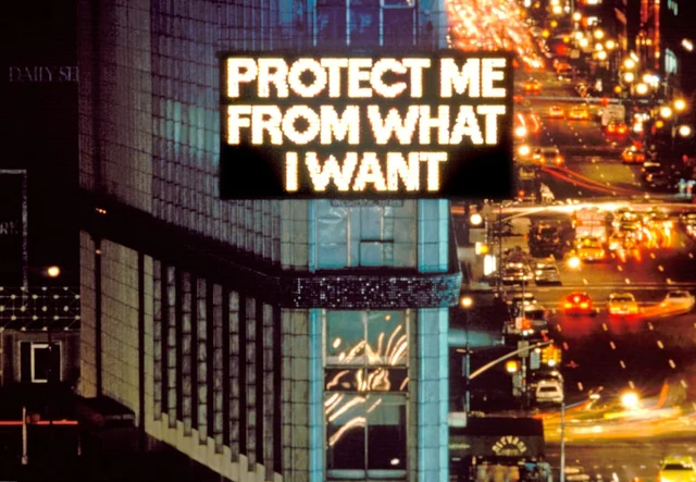 夜间俯拍时代广场，一栋大楼外墙的电子牌上写着 PROTECT ME FROM WHAT I WANT，下方是车流拉出的光带