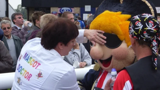 A woman in a white coat lettered "MASCOT DOCTOR" bent over a pirate mascot in a crowd