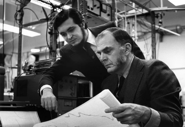 Two men in suits lean over a printed chart in a laboratory full of equipment