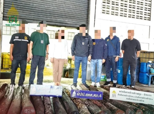 Seven people in plain clothes standing behind a row of long dark logs laid on the ground, with the Thai Customs emblem in the corner of the photograph
