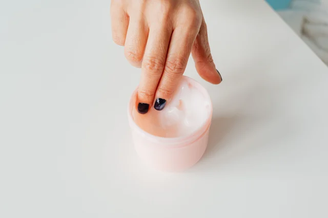 Close-up of a hand scooping thick cream from a glass jar — softly lit, warm neutral tones, illustrative of a cushiony moisturizer texture