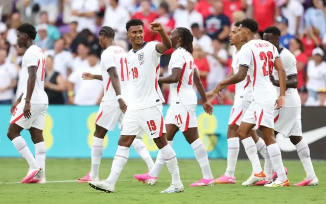 England players celebrating after a goal, in white kit, against a blurred stadium background.