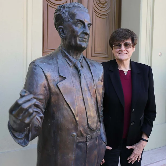 Karikó standing beside a bronze statue of a man in a suit, outside a university building