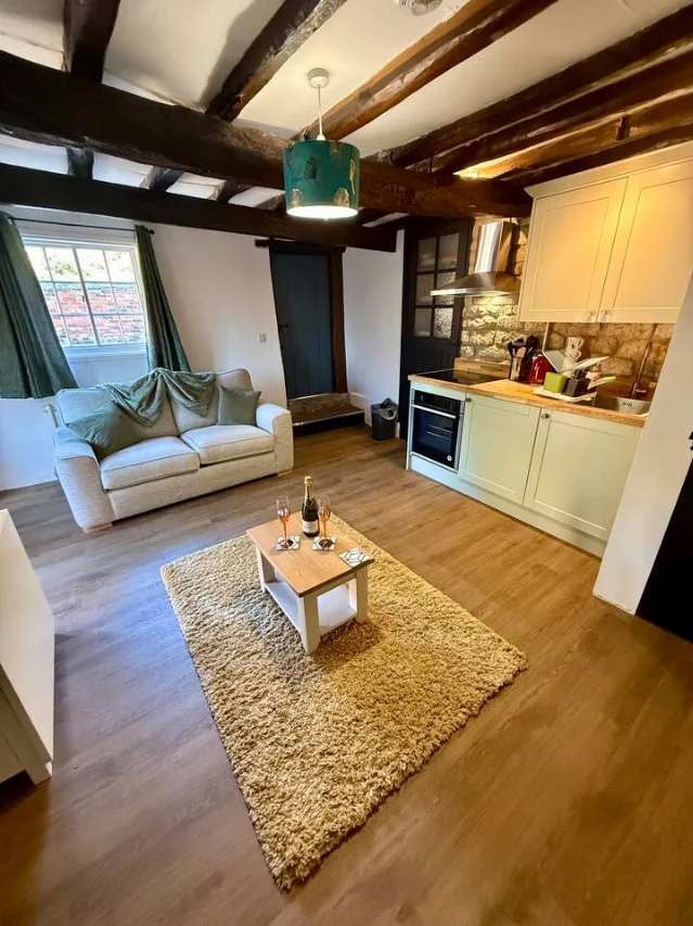 An open-plan lounge and kitchen under heavy original beams, with a sofa, a shag rug and a small dining table set with a bottle of wine