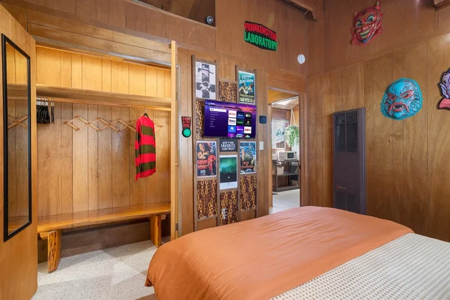 A wood-panelled bedroom with an orange-covered bed, a wall-mounted TV, framed horror movie posters, a Frankenstein Laboratory sign and rubber monster masks on the wall