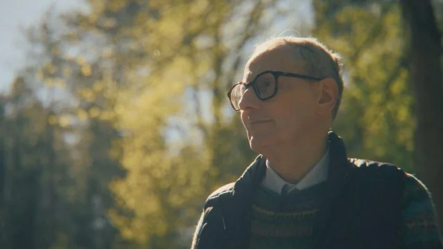 A man in dark-rimmed glasses and a knitted jumper looking up, with sunlit woodland behind him