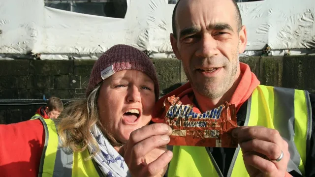 Two volunteers in high-visibility jackets holding up a crumpled, weathered Marathon chocolate wrapper between them