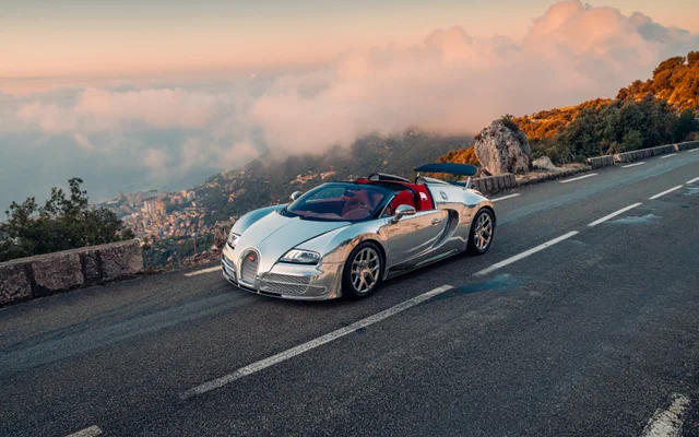 A mirror-finish Bugatti Veyron Grand Sport convertible with a red interior on a mountain road, a coastal city far below and clouds beyond