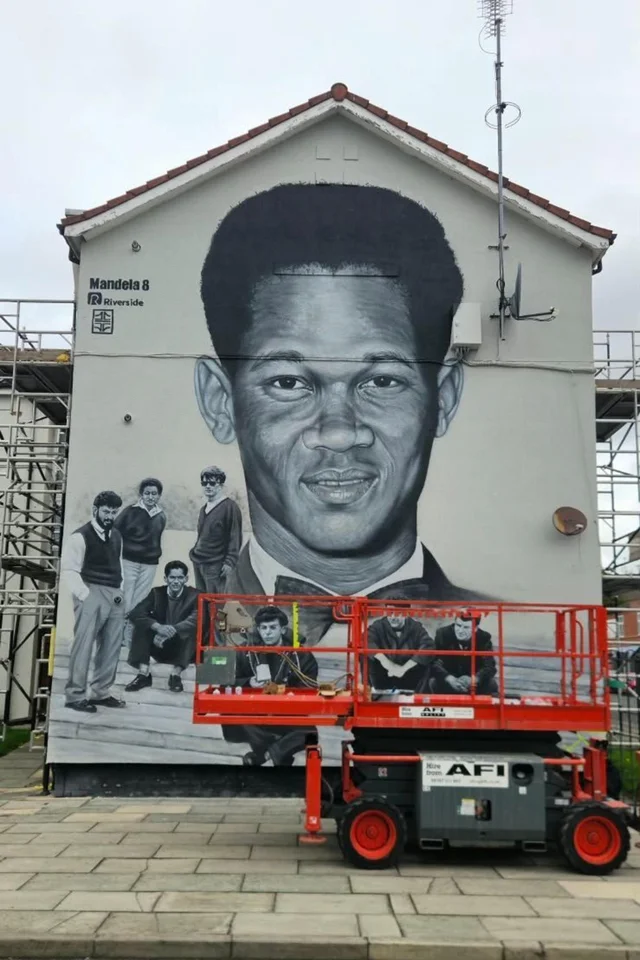 a tall terraced gable end painted with a large black-and-white portrait above a smaller painted group photograph, a red scissor lift standing in front