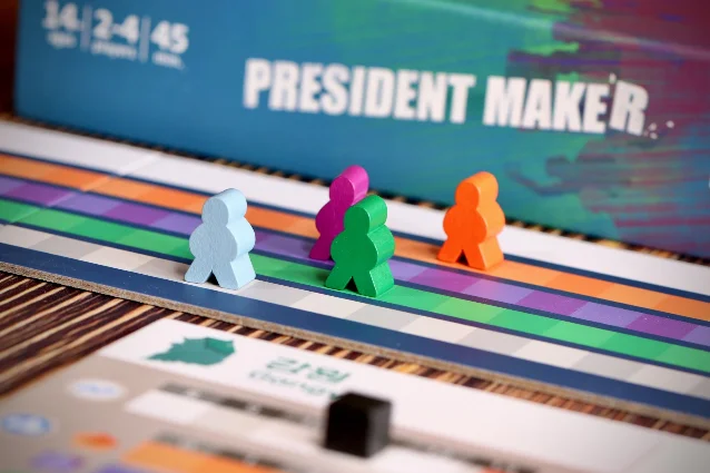 Four candidate meeples standing on the coloured support tracks of President Maker, with the box and a regional board behind them