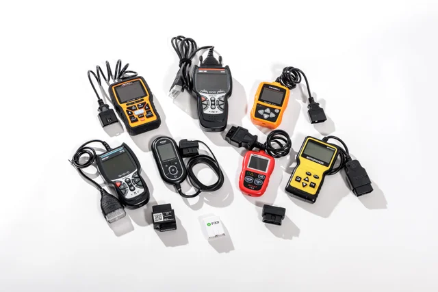 Nine OBD-II scanners laid out on a white surface, ranging from small handheld readers with coiled cords to plug-in Bluetooth adapters with no screen