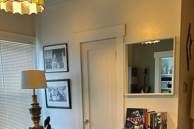 A living room corner with framed black-and-white photographs, a table lamp, a mirror reflecting an acoustic guitar, and a shelf of Nirvana books and records
